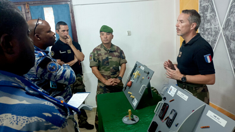 Un gendarme, vêtu d'un tee-shirt noir et d'un pantalon de treillis kaki, s'adresse à deux personnels de la gendarmerie territoriale ivoirienne, vêtus d'un treillis beige et bleu. Devant eux, plusieurs dispositifs d'IED disposés sur des présentoirs gris, posés sur une table recouverte d'une nappe verte. Au second plan un second gendarme et un militaire du 43e Bataillon d'infanterie de marine.