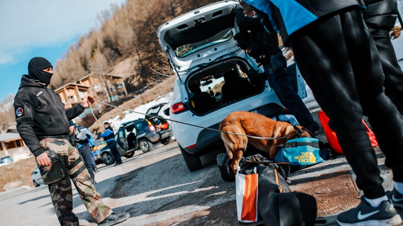 Un maître de chien tient par la laisse son malinois en train de fouiller dans des sacs au pied de l'arrière d'un véhicule. a droite, les jambes et pied d'une persone en surv^tement noir. A gauche au fond, des véhciules de gendarmerie et des gendarmes, en hauteur la montagne et des chalets