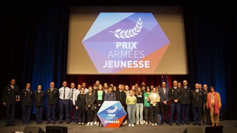 Agrandir l'image Photo de groupe avec une classe de collégiens, de part et d'autres des gendarmes en vareuse et pantalon droit bleu; sur une estrade pour la remise du prix Armées-jeunesse. Au fond sur un écran, le logo du prix Armées-Jeunesse.