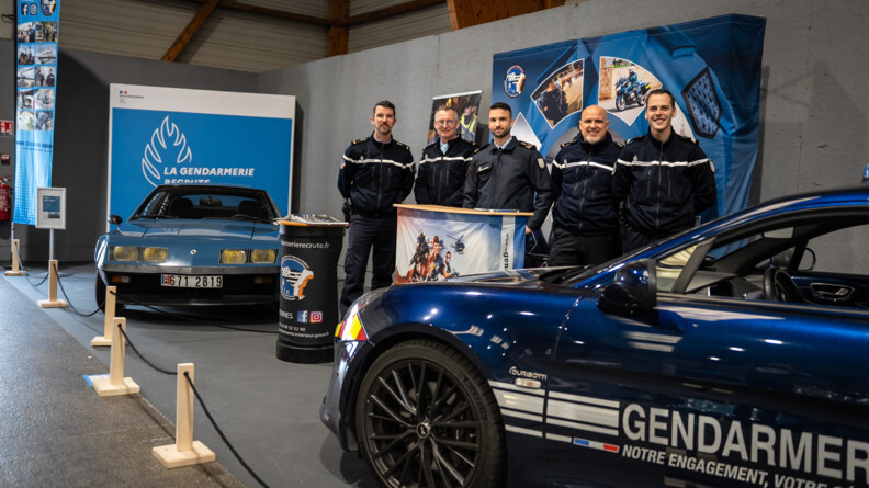 Sur un stand de la gendarmerie dans une exposition, deux alpine, une ancienne et une récente, sont exposées. Cinq gendarmes, souriant, posent derrière le stand