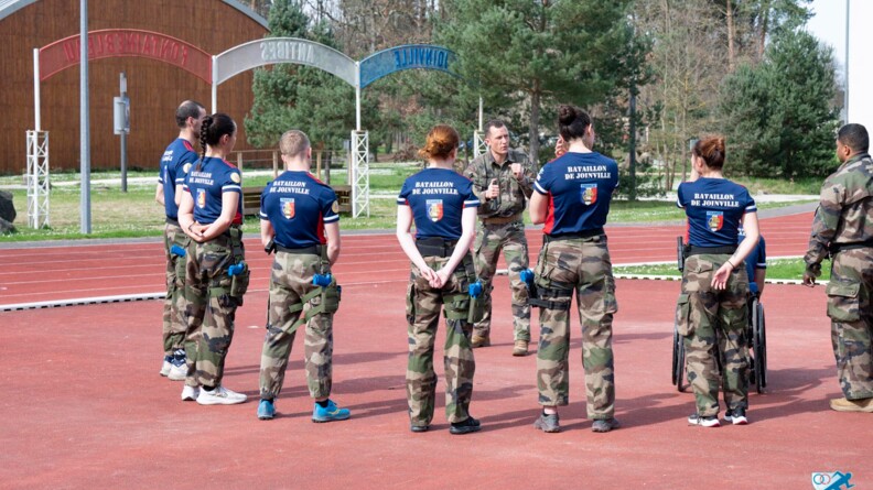 Huit membres de l'armée de champion de dos, dont un dont on devine les roues du fauteuil, en bas de treillis et t-shirt bleu siglé en blanc "Bataillon de Joinville" regardent un homme en treillis expliquant le fonctionnement d'u nbâton téléscopique. Au fond des épineux, un bâtiment couleurs bois et trois arches, une bleu, une bland, une rouge