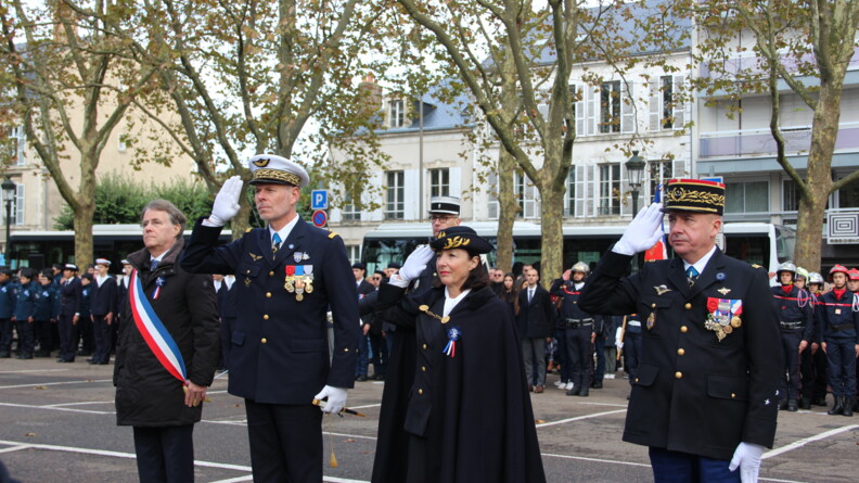 Sur un parking extérieur,  au sol gris délimité par des blandes blanches, se tiennent trois militaires (deux hommes et une femmes) effectuant un salut, aux côtés du maire, qui portent une écharpe tricolore sur le devant du corps. Derrière eux, se tiennent des personnes, civiles et militaires. On aperçoit les façades des immeubles ainsi que les arbres ayant partiellement perdu leurs feuilles.