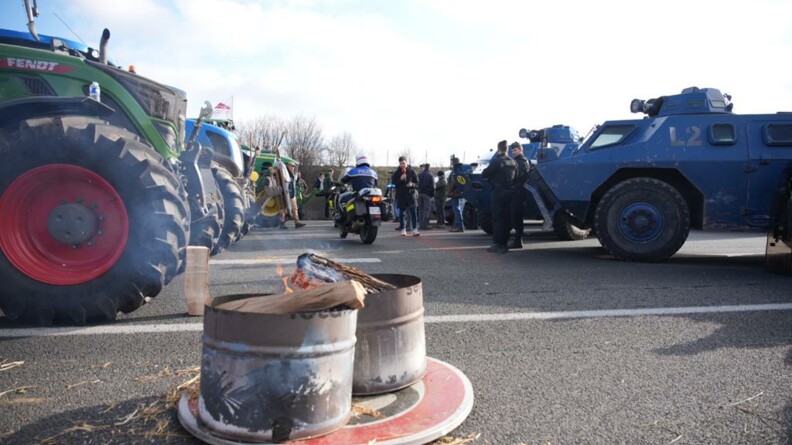 Sur le côté gauche de la photo se trouve des tracteurs, en face, sur le côté droit, leur font face des véhicules blindés de la gendarmerie. Au milieu, un feu a été allumé dans de grands bidons métalliques et un motard de gendarmerie passe.