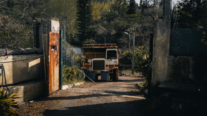 Devant un portail ouvert, un camion blanc rouge et roull&eacute; est stationn&eacute; derri&egrave;re des poubelles. Derri&egrave;re, la v&eacute;g&eacute;tation et les montagnes