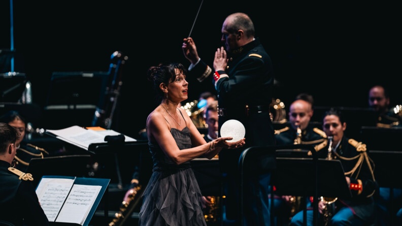 Une femme tenant un globe lumineux dans les mains avec une robe longue grise sur une scène au milieu de l'orchestre de la garde républicaine