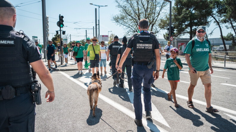 Une patrouille cynophile REXPEMO vue de dos traverse le flux de supporters arrivant face &agrave; elle. Au premier plan &agrave; gauche, un autre gendarme vu de dos.