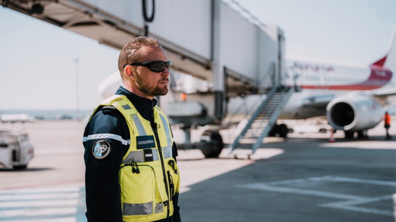 Un gendarme de la brigade de gendarmerie des transports aériens, portant un gilet jaune, par-dessus son uniforme bleu marine, ainsi que des lunettes de soleil noires, se tient debout, de biais. L'homme regarde droit devant lui. Derrière lui, en arrière plan, on aperçoit un avion de ligne blanc et rouge, stationné à proximité immédiate d'une passerelle