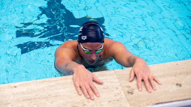 Une nageuse revêtant un bonnet bleu marine ainsi que des lunettes de nage, récupère, au bord du bassin, les mains posées sur le rebord de la piscine