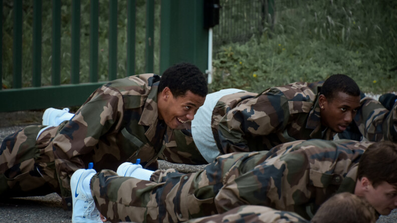 Quatre hommes en treillis kaki de trois-quarts face, en train de faire des pompes.