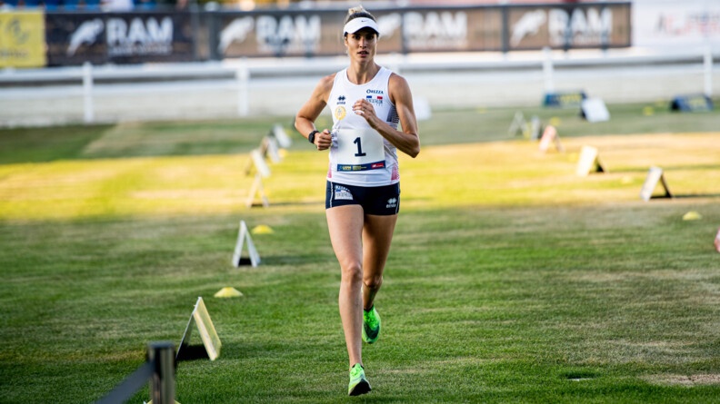 Une femme en tenue de sport avec une casquette en train de courir dans l'herbe, son chemin est démarqué par des plots jaunes et des panneaux posés sur le sol