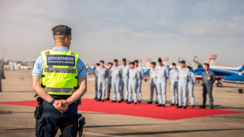 De dos à gauche,un gendarme de la GTA, mains dans le dos, avec une chasuble jaune. Au second plan, flou, les pilotes de la patrouille de France aligné sur un tapis rouge, sur le tarmac, et leurs avions