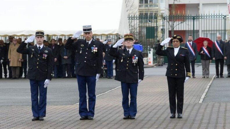 Quatre personnes, autorités civiles et militaires, au garde-à-vous. Dans le fond, un parterre d'invités, dont trois sur la droite arborent une écharpe tricolore.