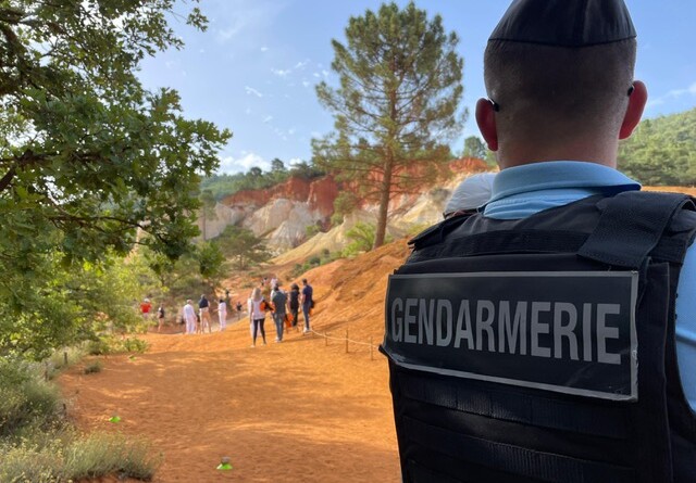Un gendarme vu de dos, avec la mention GENDARMERIE sur son gilet pare-balles, est posté au bord d'un chemin couleur ocre, bordé de quelques arbres. Au loin, plusieurs presonnes marchent ou sont statiques.