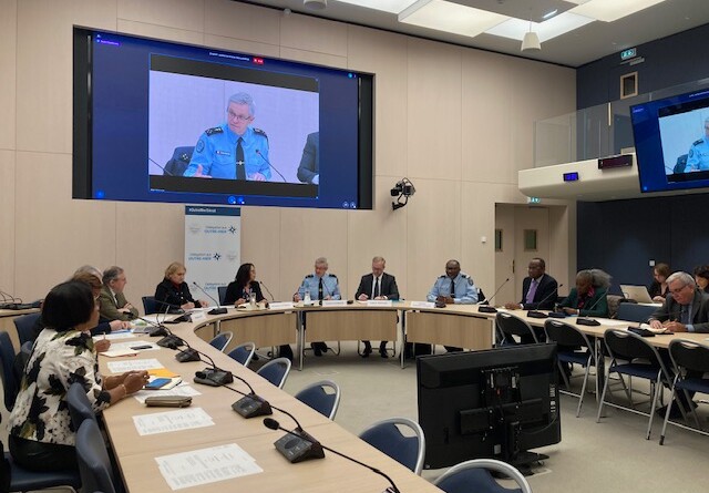 Agrandir l'image Dans une vaste salle aux murs beiges, des hommes et des femmes, civils et gendarmes en uniformes, sont assis autour d'une grande table circulaire, avec des micros devant chacun d'eux. Au mur est accroché un grand écran télé, sur lequel apparait un homme en uniforme.