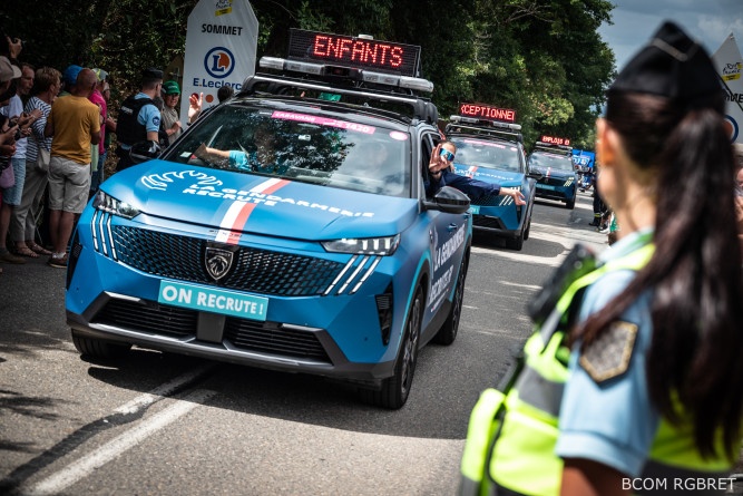 Agrandir l'image Une gendarme en uniforme, avec un gilet tactique jaune fluo et une queue de cheval, vue de trois-quart dos, est positionnée sur le bord de la route, tandis que les véhicules gendarmerie de la caravane du Tour de France passent devant elle. De l'autre côté de la chaussée sont massées de nombreuses personnes.
