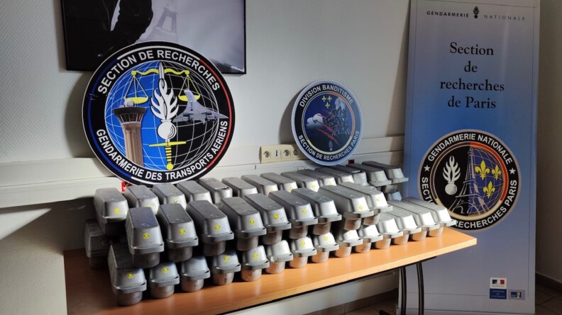 Sur une table sont alignées une cinquantaine de batteries grises de trottinettes, surmontées du logo de de la section de recherches des transports aériens, celui de la division banditisme de la section de recherches (S.R.) de Paris, et à droite un kakémono de la section de recherches de Paris. Le tout contre un mur blanc.