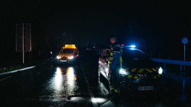 Sous la pluie et de nuit, un gendarme en tenue attente le passage du convoi devant sa voiture. En arrière plan, une voiture annonçant "Convoi exceptionnel" est entrain d'arriver.