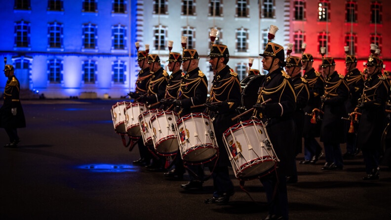 Agrandir l'image La musique de la garde republicaine, vue de profil gauche, avec 6 tambours alignés et les trompettes alignées au second rang. EN fond, l'etat major de l'EOGN illuminé en bleu blanc rouge