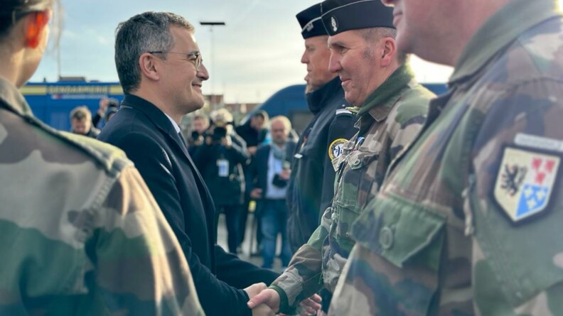 G&eacute;rald Darmanin, debout, de biais, v&ecirc;tu d'un manteau sombre, souriant, serre la main d'un militaire en tenue kakie. Ils sont en ext&eacute;rieur. Le ciel est bleu et nuageux &agrave; la fois.