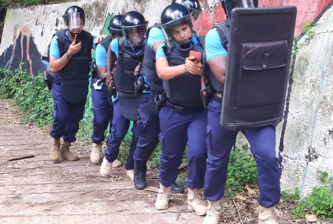 Colonne de six policiers seychellois progressant le long d'un mur, derri&egrave;re un bouclier.