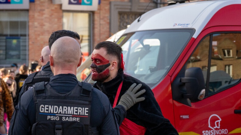 Photo de deux gendarmes, de dos, dans la rue, parlant avec un carnavaleux, au visage maquill&eacute; en noir et rouge, lors du carnaval de Bergues, devant un camion de pompiers