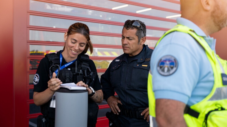 A gauche, une gendarme avec un calepin appuyé sur un petit pilier gris. A sa droite, un gendarme, les mains sur les hanches, regarde son calepin. A droite, flou, au premier plan, un gendarme de la GTA avec sa chasuble. Le fond est une porte rouge.