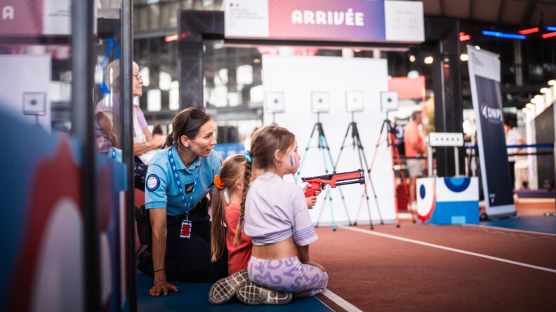 Agrandir l'image Une enfant blonde, que l'on voit de profil, tire avec une carabine orange sur un stand de tir. A côté d'elle se tient une autre fillette, blonde également, avec le drapeau français imprimé sur la joue, ainsi qu'une femme gendarme qui la guide dans ses mouvements.