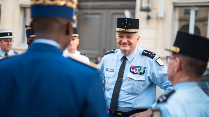 Agrandir l'image De face, se tient debout un homme, le directeur général de la gendarmerie nationale, en tenue.. deux hommes, que l'on voit se dos, lui fony face : un gendarme congolais et une autorité de la gendarmerie française. Derrière eux se trouve un bâtiment en pierre, dont on distingue une grande porte noire et une lanterne