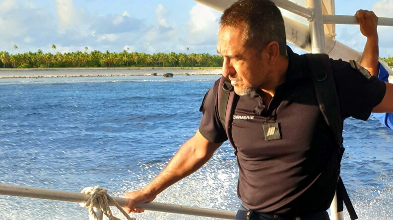 Sur un bateau, l'adjudant Teihotua se tient à une rembarde. Derrière lui, la mer et une île avec des palmiers.