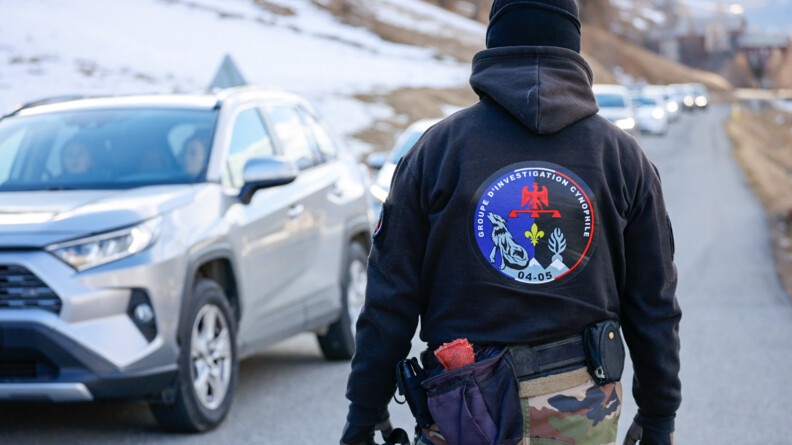 De dos un gendarme en treillis et haut noir du Groupe d'investigation cynophile devant une file de véhicules sur une route en bord de montagnes
