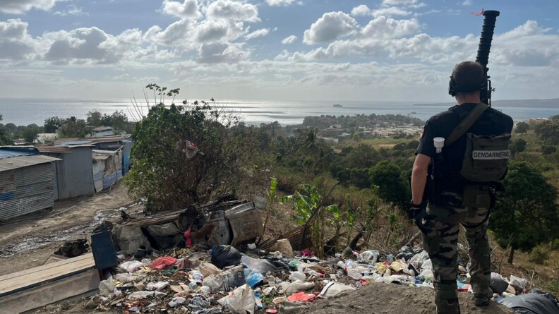 Un gendarme mobile, pantalon de treillis kaki et tee-shirt noir, de dos. Devant lui un tas d'immondices, &agrave; gauche des habitats en t&ocirc;le. Dans le fond la verdure et l'oc&eacute;an.