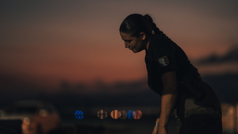 Une femme gendarme se tient de bout, le corps légèrement incliné en avant. La scène se déroule la nuit.