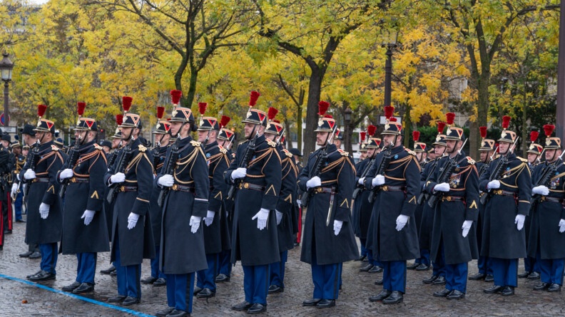 Sur un sol pavé, devant des arbres feuillus aux couleurs de l'automne, se tiennent des militaires de la Garde républicaine. Vêtus d'un long manteau noir, d'un pantalon bleu marine et du traditionnel chapeau noir à plumes rouges, ils tiennent leur fusil serré contre leur poitrine.