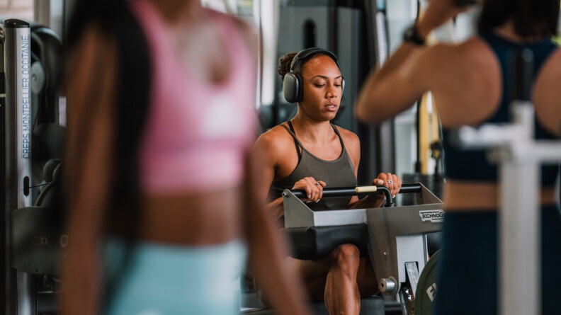 Une sportive vêtue d'une tenue grise, effectue un exercice sur un appareil de musculation. Elle porte un casque sur les oreilles.