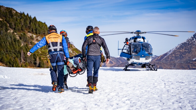 Quatre gendarmes secouristes portent un individu dans un brancard. Au fond de l'image, on voit l'hélicoptère qui les attends.