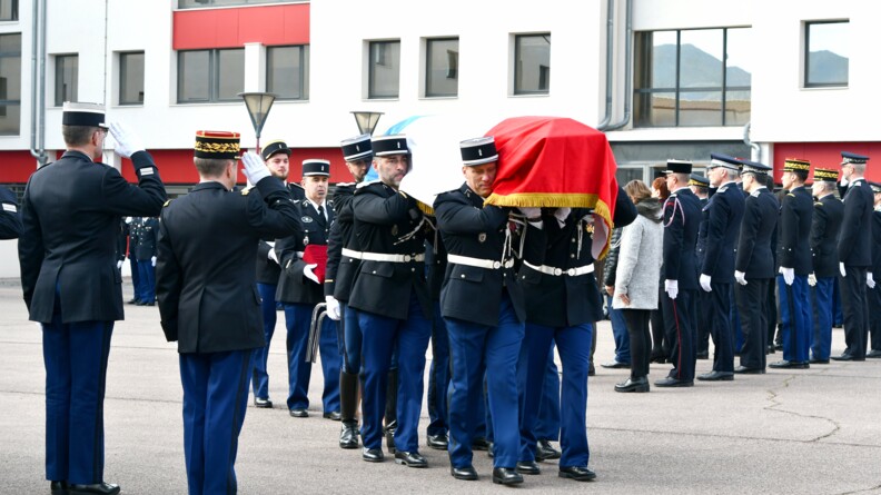 Au premier rang, de dos, deux gendarmes au garde-à-vous. Devant eux passe un cercueil drapé du drapeau tricolore, porté par six gendarmes. En arrière-plan, à droite, deux rangées de militaires et de civils debout, vus de dos.