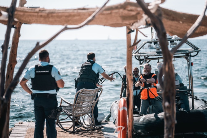 Agrandir l'image Deux gendarmes se tiennent deboit sur un ponton, tournant le dos à l'objectif. Alors qu'une petite embarcation s'apprète à accoster, l'homme reçoit dans ses mains la corde jetée par l'un des occupants du bateau.