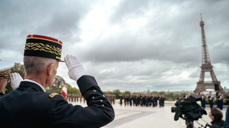 Un haut gradé, que l'on aperçoit de dos, se tient debout sur l'esplanade du trocadéro, face à la Tour Eiffel, revếtant les anneaux olympiques. L'homme effectue un salut militaire, en direction de militaires se tenant debout face à lui, et dont l'image est floutée.