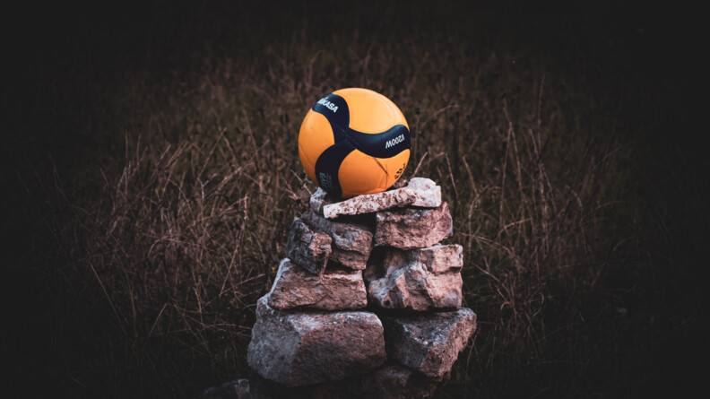 Au centre de l'image, un ballon de volley noir et jaune-orangé, posé sur un monticule de pierres, au milieu d'un champ.