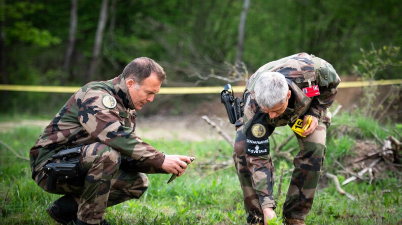 Deux gendarmes de la prévôté en treillis, accroupis, en train de faire des constatations en déposant des marqueurs jaunes sur l'herbe