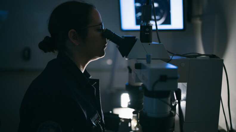 Dans une pièce très sombre, sans éclairage, on aperçoit une femme de profil, vêtue d'un uniforme bleu marine. Elle porte des lunettes, et un chignon bas. Elle regarde dans la lunette d'un macroscope. dans le fond de la pièce, un écran blanc lumineux projette une image. Il s'agit d'une forme circulaire de couleur blanche sur fond noir.