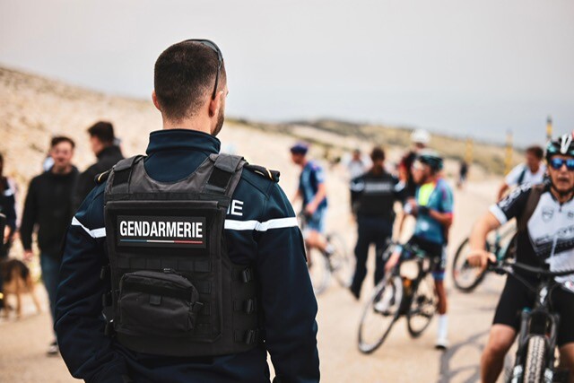 Au premier plan, un gendarme de dos, portant un gilet pare-balles siglé GENDARMERIE, par dessus une polaire. Au second plan, plusieurs cyclistes, pied à terre, et des gens à pied, un autre gendarme plus loin, dans un paysage minéral (Mont Ventoux), sous un ciel gris.