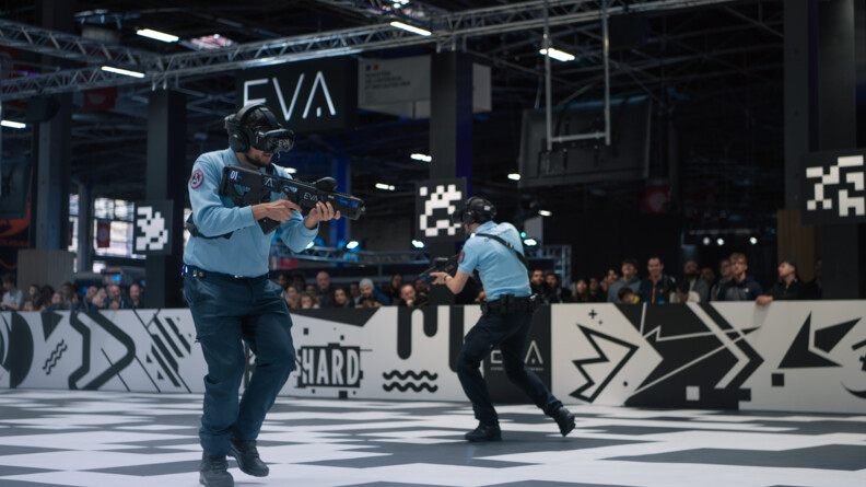 Dans un grand hall, deux gendarmes jouent sur un sol noir et blanc. Ils avancent debout, tenant une arme de jeu devant eux. Tout autour, des spectateurs assistent à la scène.