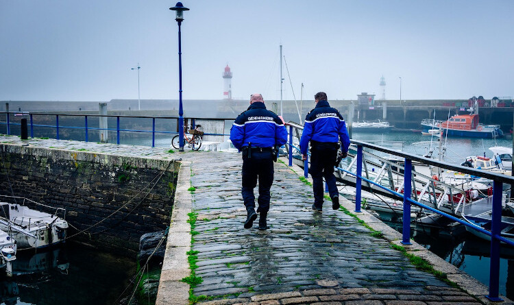 Agrandir l'image Deux gendarmes de dos en patrouille le long de la jetée d'un port. De part et d'autre, sous un ciel brumeux, des petits bateaux amarrés. Au fond de la scène apparaissent les tours de deux phares