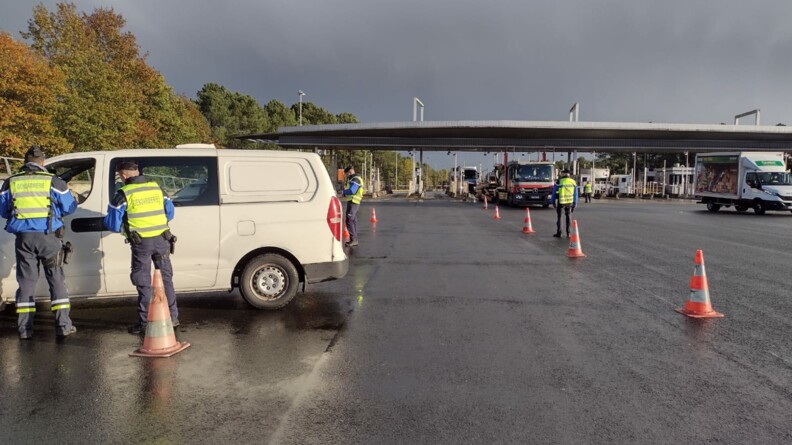 Agrandir l'image Au premier plan, à gauche, deux gendarmes, vêtus d'une chasuble jaune, en train de contrôler le conducteur d'une camionnette blanche. En arrière-plan, une barrière de péage et plusieurs autres gedarmes scrutant le flux routier.