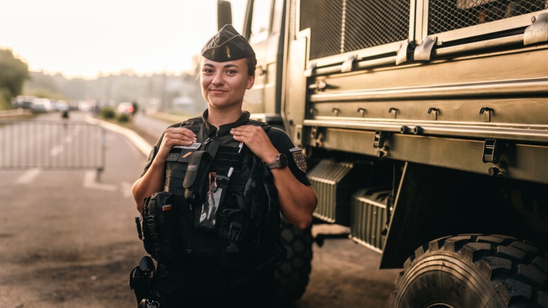 Une jeune femme gendarme, vêtue d'une tenue noire et d'un gilet par-balles, se tient debout, sur une large avenue, devant un véhicule blindé de couleur kakie.