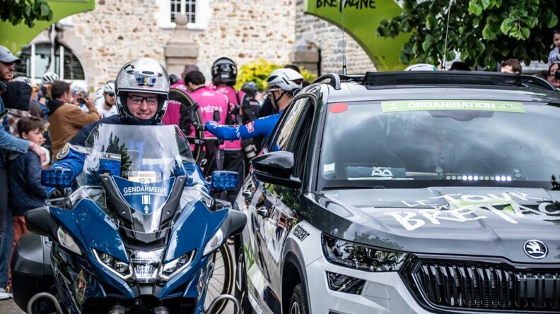 Au premier plan, un motocycliste de la garde républicaine à côté d'un véhicule de course. Au second plan, des coureurs et des spectateurs.