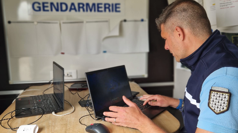 Un gendarme assis devant une table, que l'on voit de biais, tape sur le clavier de son ordinateur. derrière lui, sur le mur, on peut lire le mention "PC GENDARMERIE"
