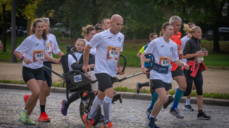 Des coureurs, vêtus d'un T-shirt blanc avec un numéro sur le ventre, courent sur une avenue pavée, en poussant le fauteuil roulant d'une jeune femme blonde, portant des tresses. On aperçoit des arbres aux feuilles vertes en arrière plan.