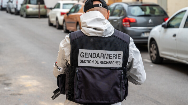 Un homme de dos avec une casquette, une parka claire et une chasuble noire avec l'inscription "gendarmerie section de recherches"